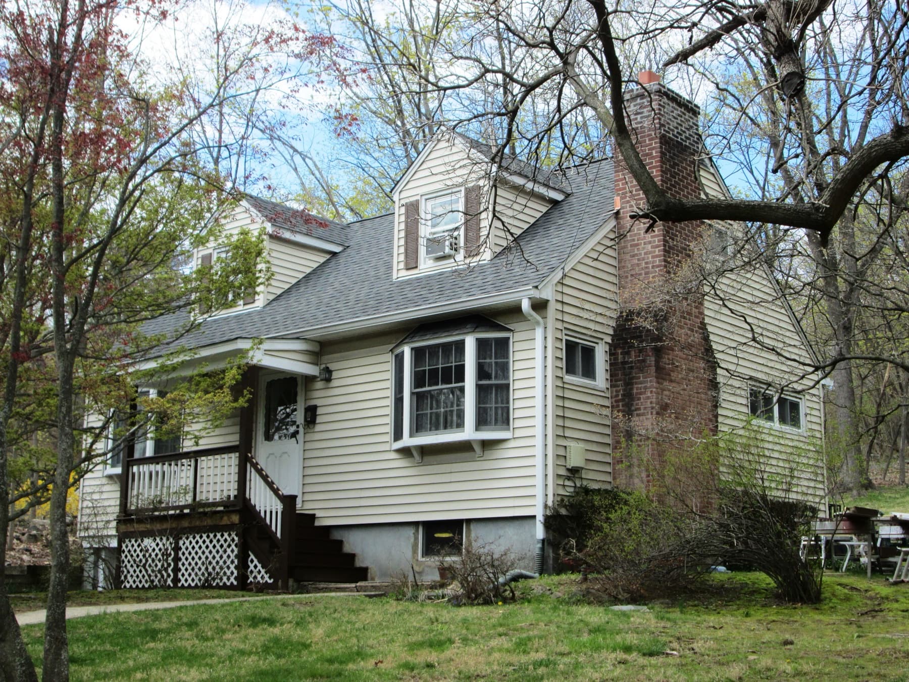 Lived-in beige Cape Cod with dormers and a brick chimney — ordinary suburban homes, not luxury estates
